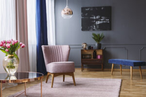 Real photo of a pink armchair standing on a rug and under a lamp in spacious living room interior, next to a table with flowers and in front of a shelf next to a grey wall with dark painting