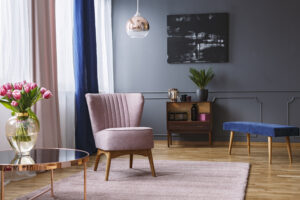 Real photo of a pink armchair standing on a rug and under a lamp in spacious living room interior, next to a table with flowers and in front of a shelf next to a grey wall with dark painting